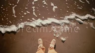 一种人的靴子的特写，站在沙质的海岸上，脚下有波浪。
